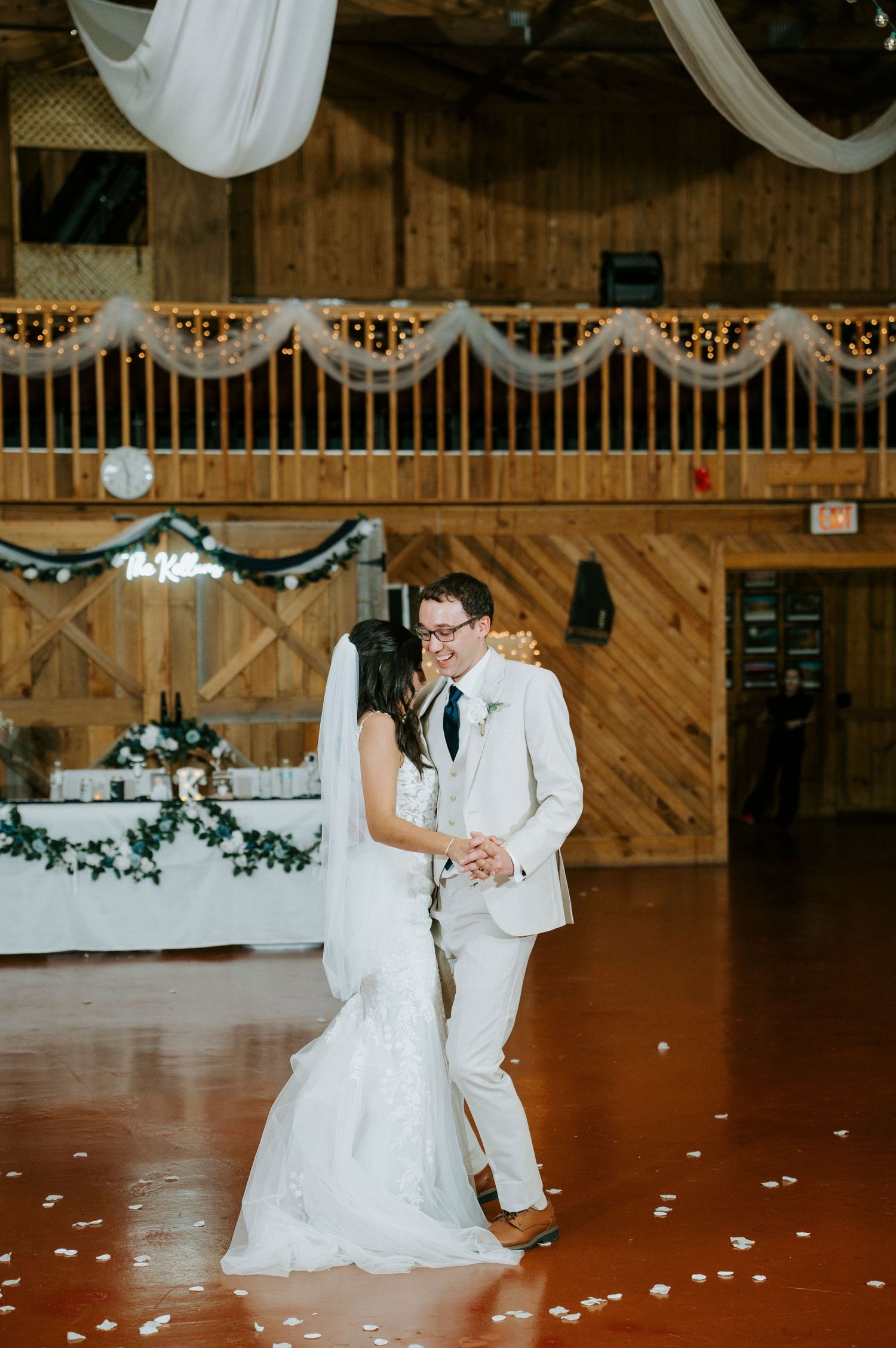 A couple shares their first dance at a rustic barn wedding reception with white draping and decorative lights.
