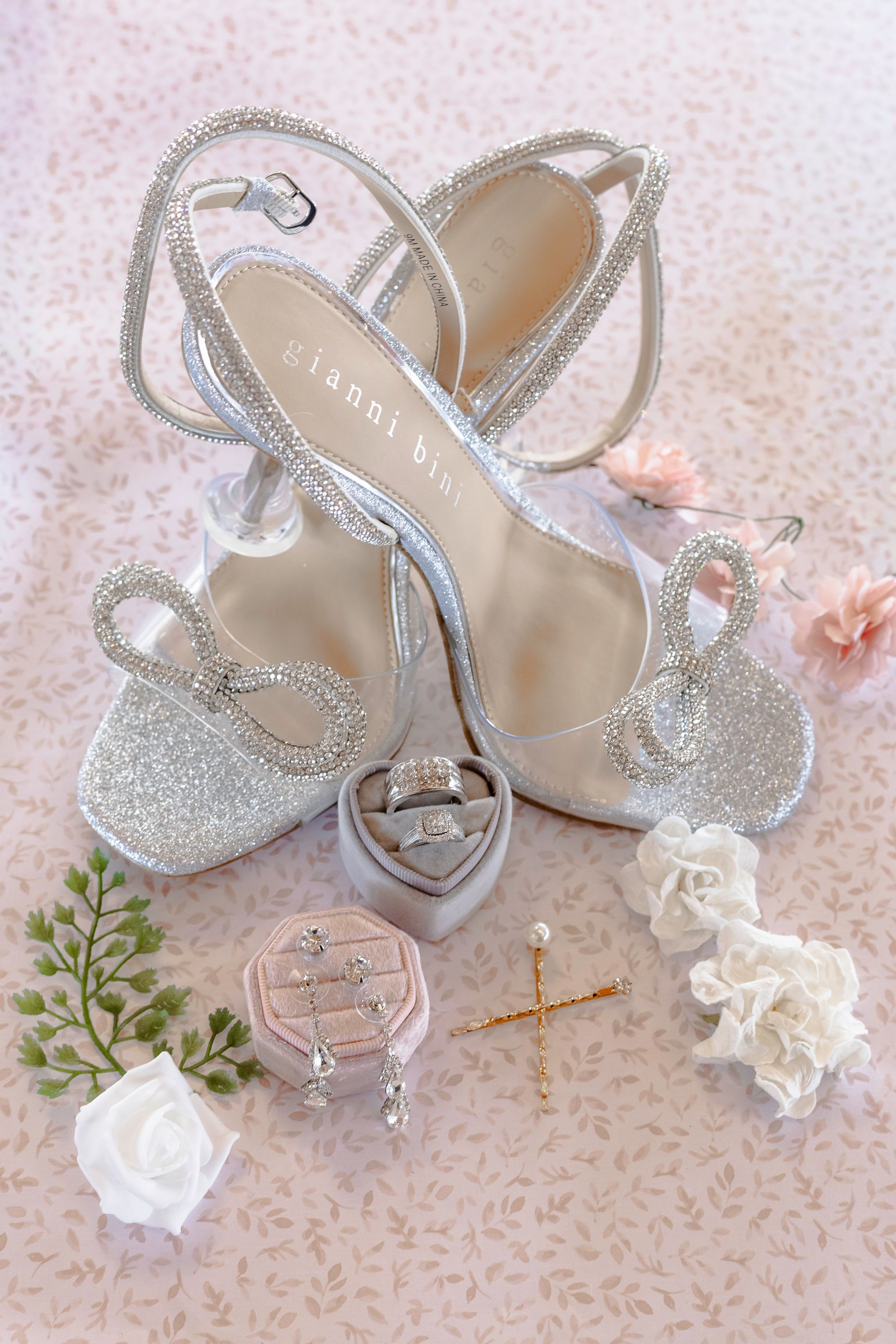 Sparkly silver wedding heels with glitter detail displayed with delicate white flowers and pink accents on lace background.