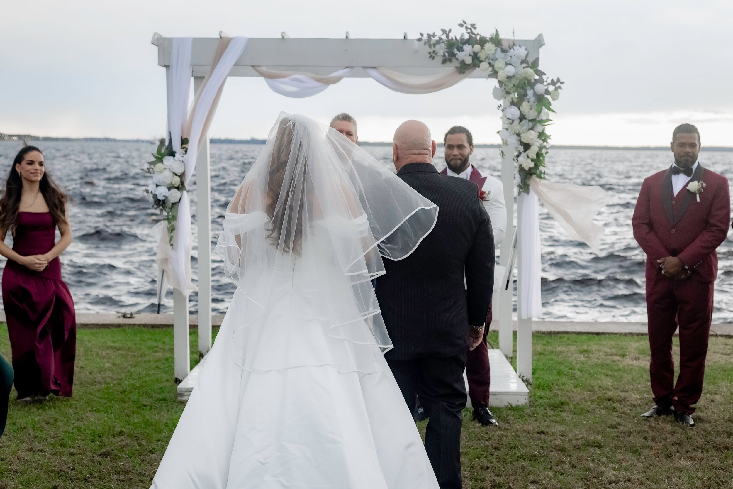 A wedding ceremony setup with white drapes and chairs by a waterfront during sunset.