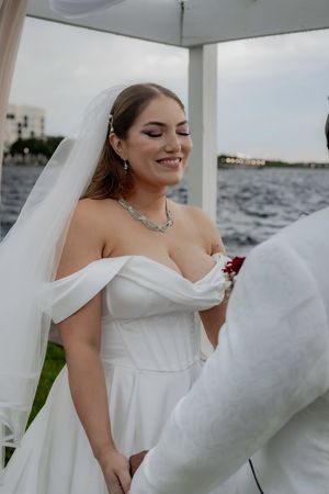 A bride in a white off-shoulder wedding dress poses by the water with her veil flowing in the breeze.