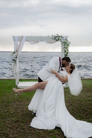 Couple shares romantic dip kiss by waterfront with flowing white wedding dress and floral arch.