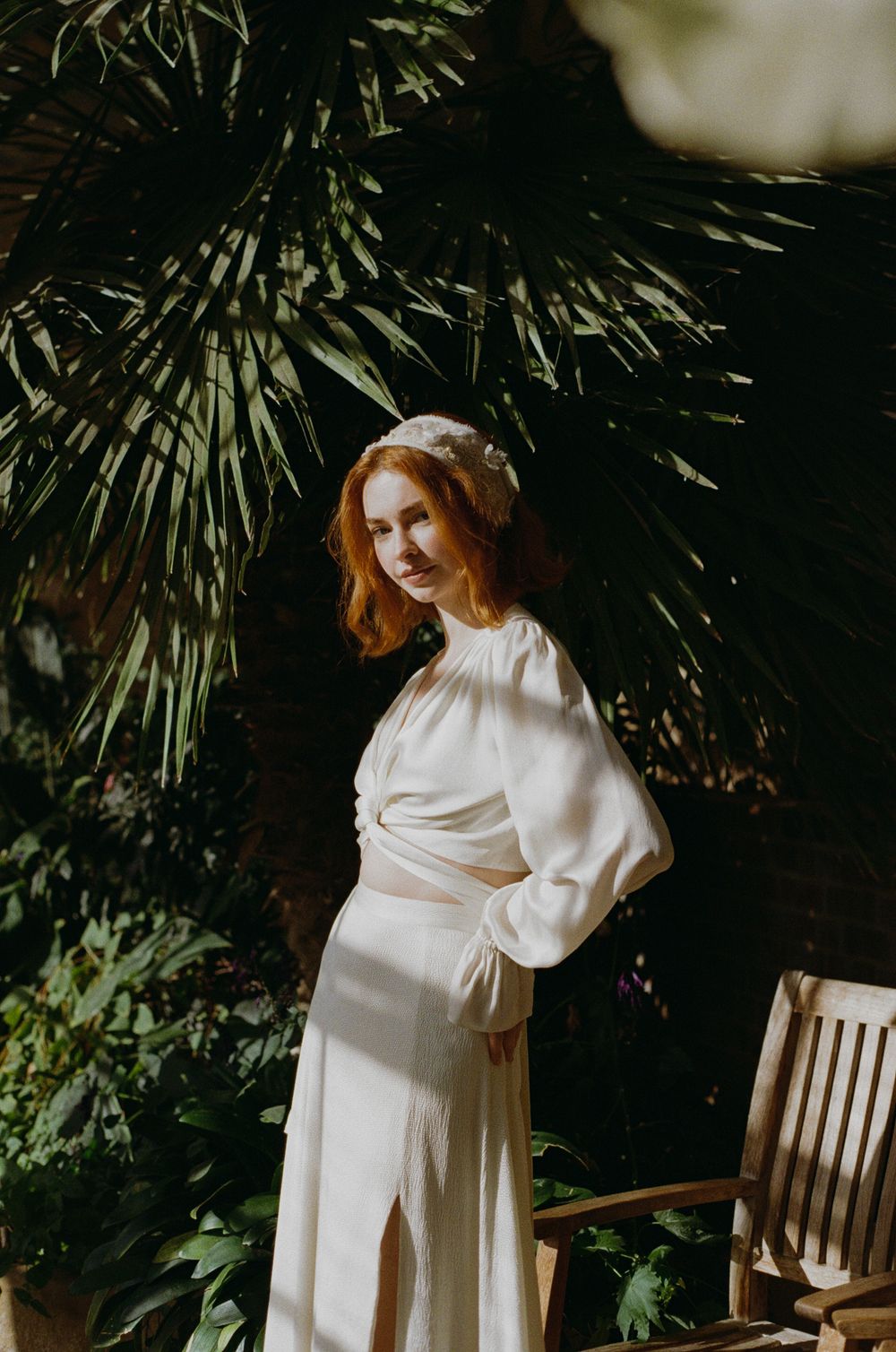 A figure in a flowing white dress poses elegantly against a backdrop of lush tropical palm fronds in a garden setting.