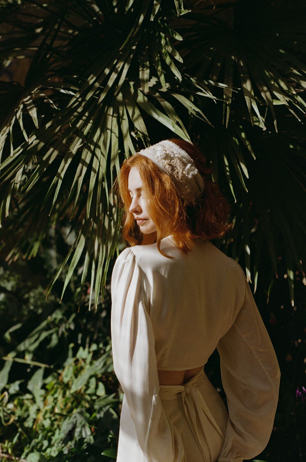 A person in a white dress stands against a backdrop of lush green foliage in dramatic sunlight.