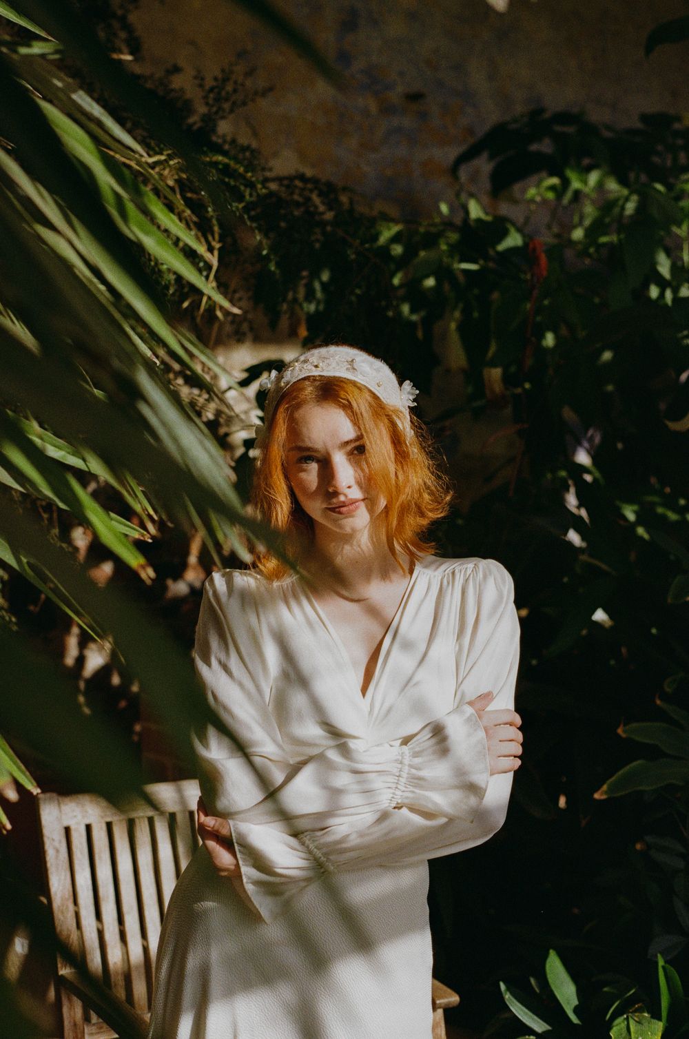 A figure in a white silk dress poses against a dark background with green foliage creating a moody garden atmosphere.