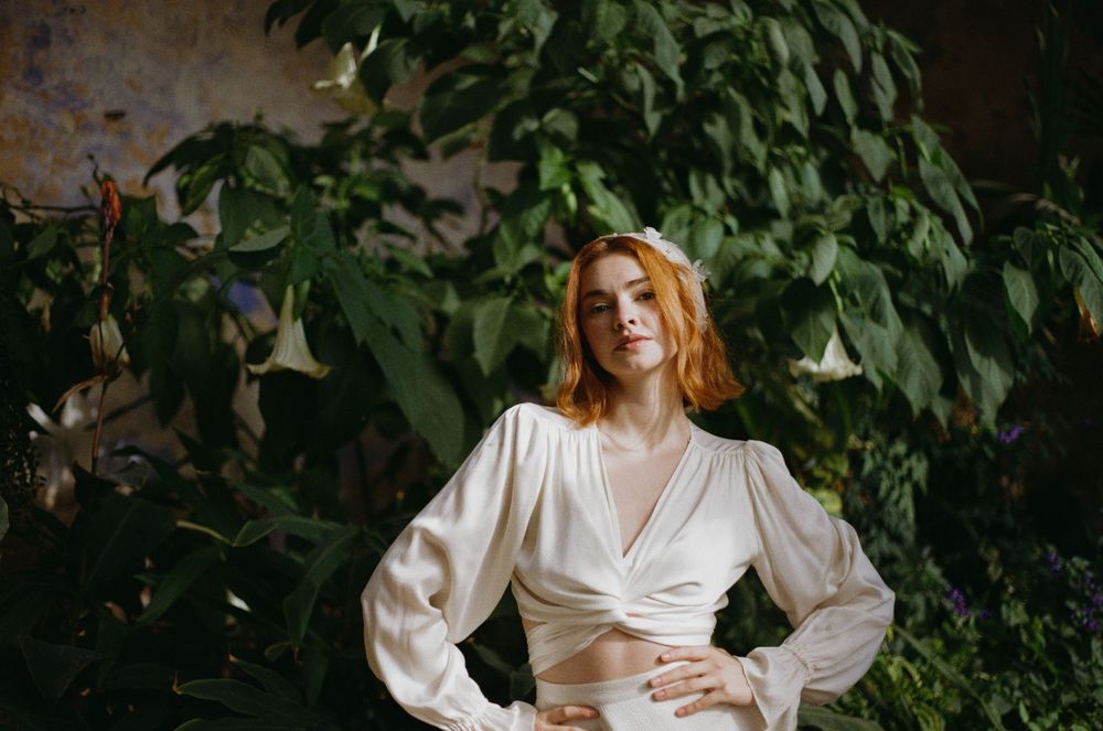 A woman in a vintage white dress poses in front of lush green foliage in an outdoor garden setting.