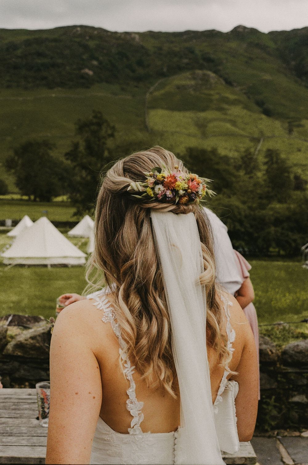 Wedding party wearing floral crowns and veils stand together against scenic green mountain backdrop.