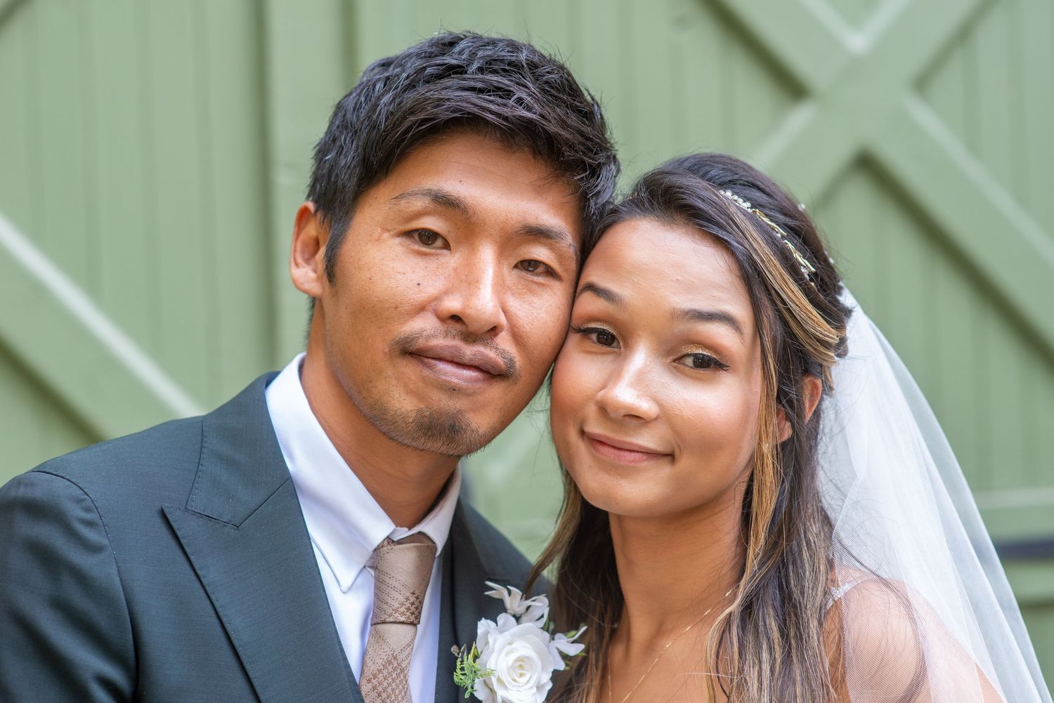 A happy couple shares a tender moment in their wedding attire against a green wall background.