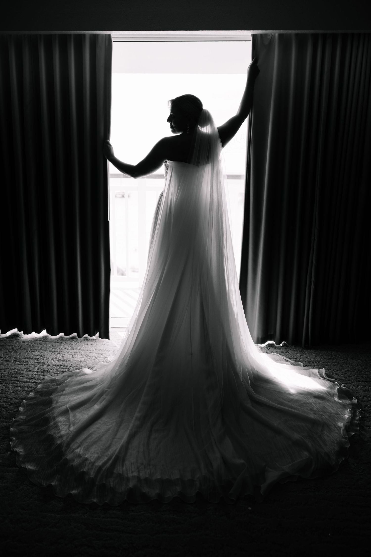 Dramatic silhouette of wedding dress against window light in black and white.