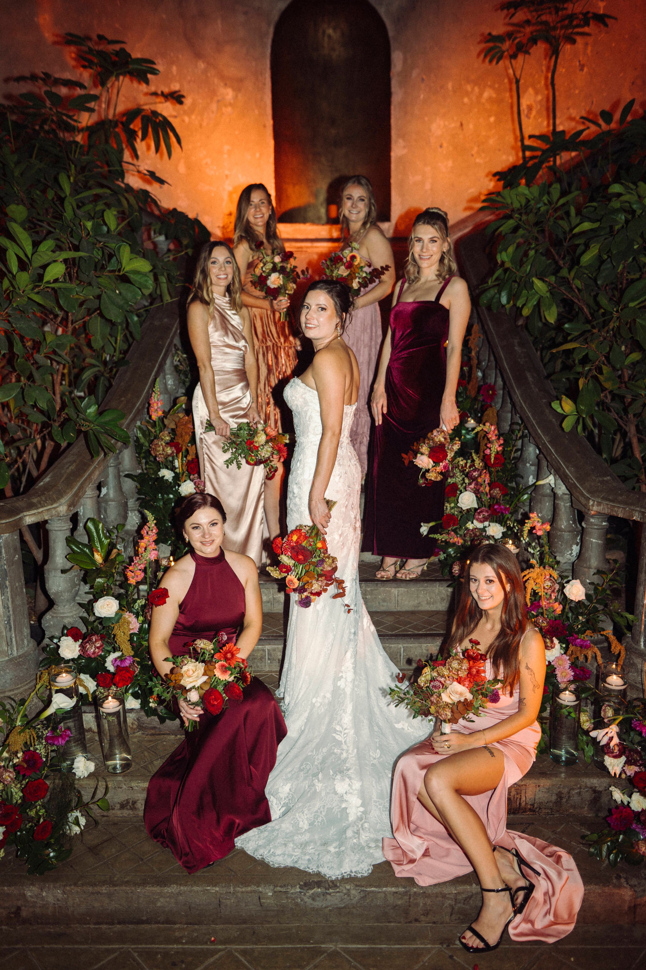 Wedding party poses on stone steps at sunset with burgundy and pink dresses and floral arrangements.