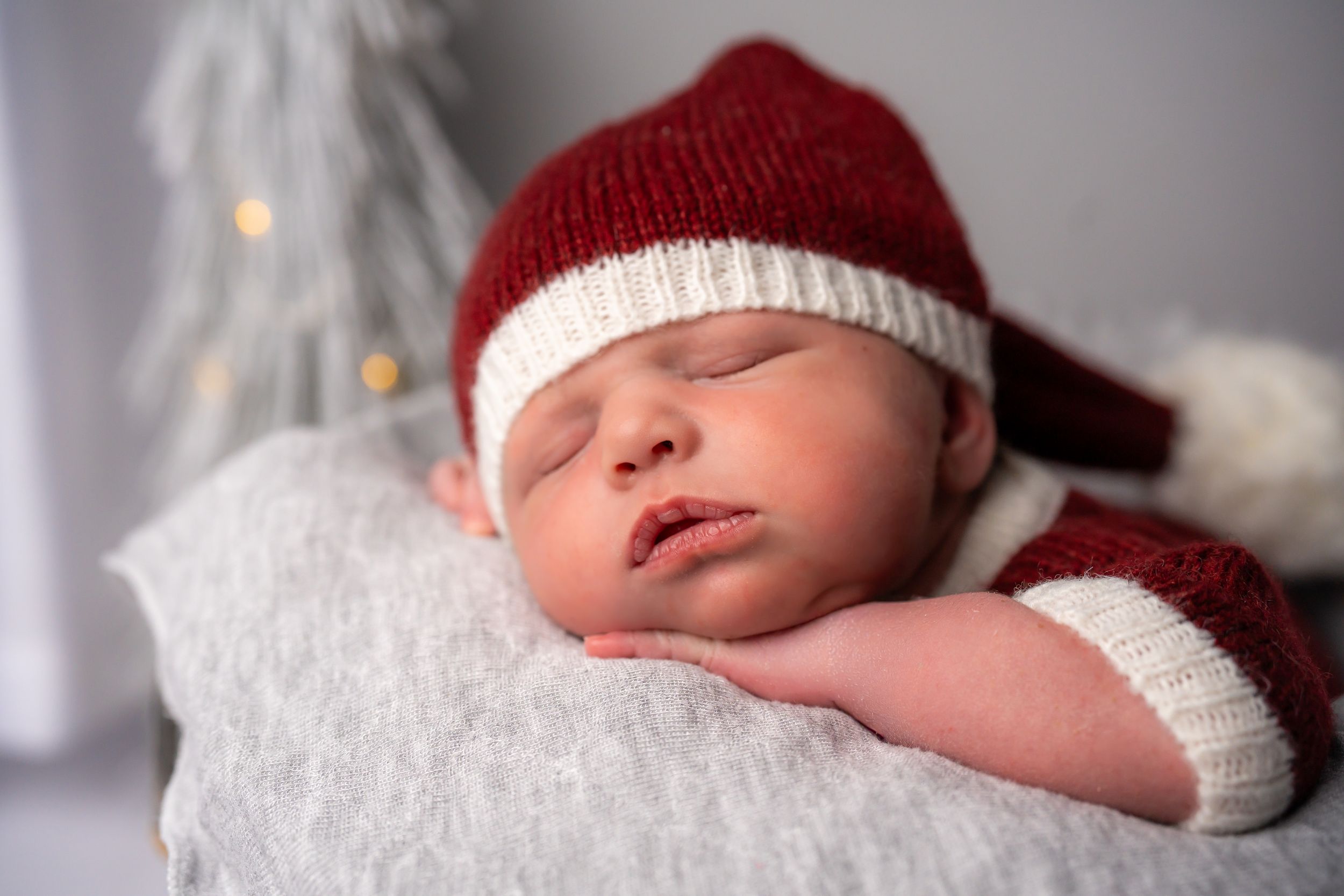 A newborn baby wearing a red knit cap rests peacefully in soft white blankets.