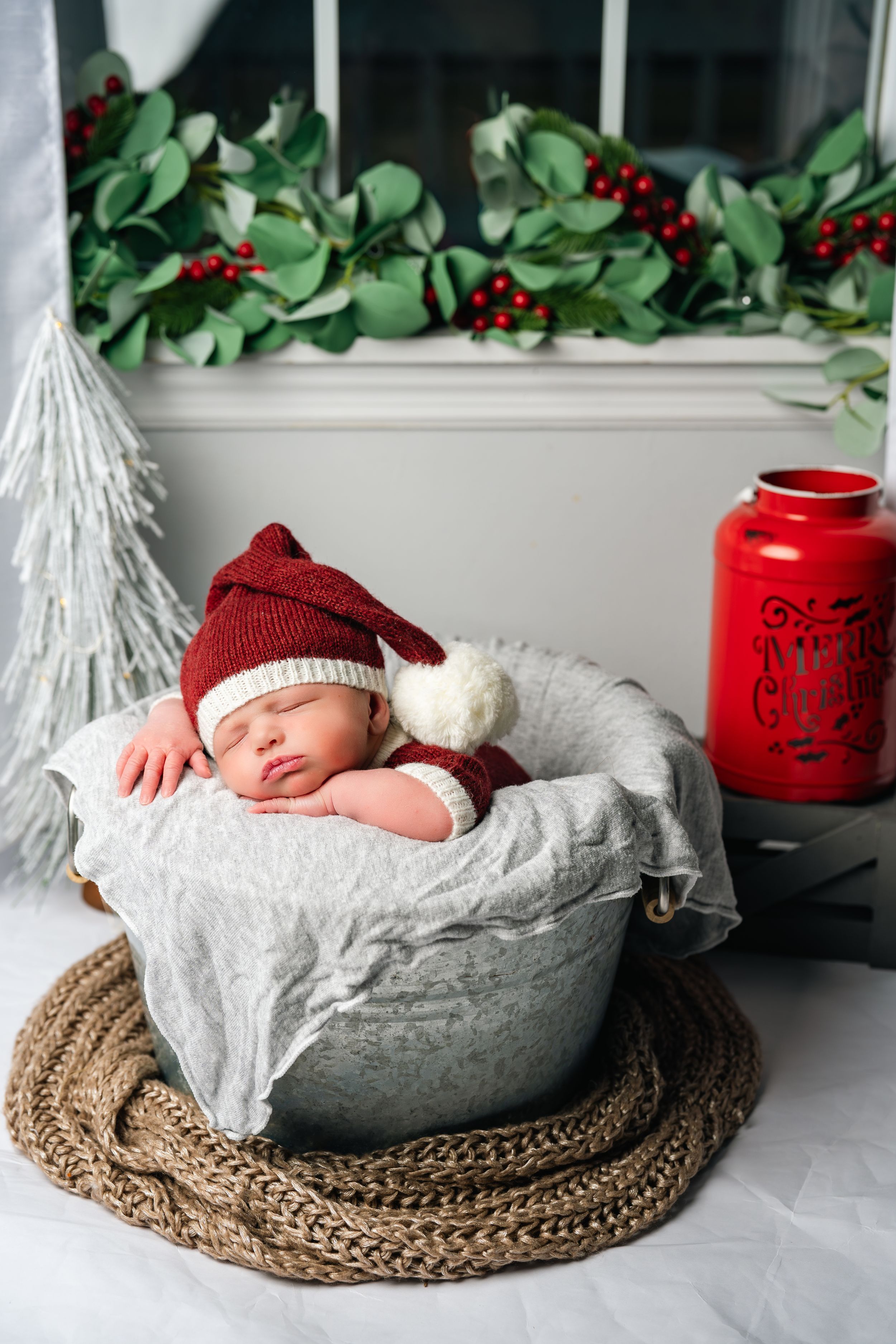 A sleeping newborn in red hat lies in a white basket on brown woven mat with festive holiday greenery and decor.