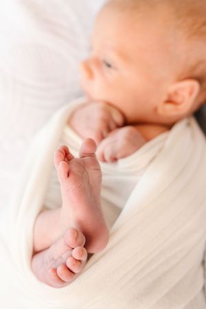 Close-up of a newborn baby wrapped in a soft white swaddle blanket, with tiny feet and toes visible in focus.
