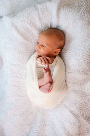 Newborn baby swaddled in white wrap lying on a textured white blanket with tiny feet peeking out.