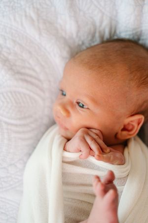 Newborn baby swaddled in white blanket with tiny hand near face, lying on white textured bedding.