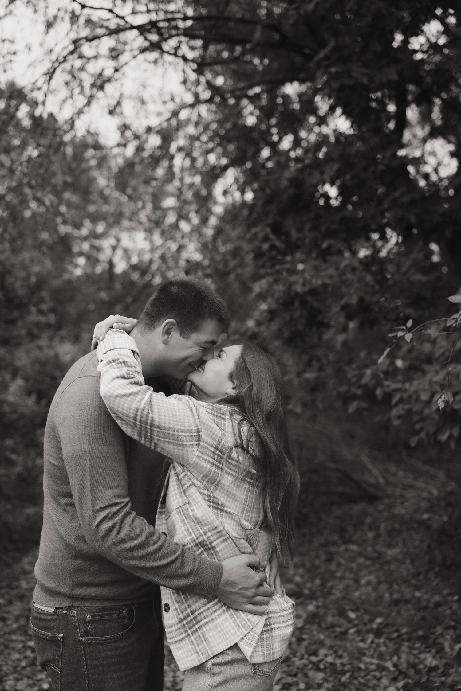 Black and white photo of a romantic embrace between a couple in casual clothes among trees.
