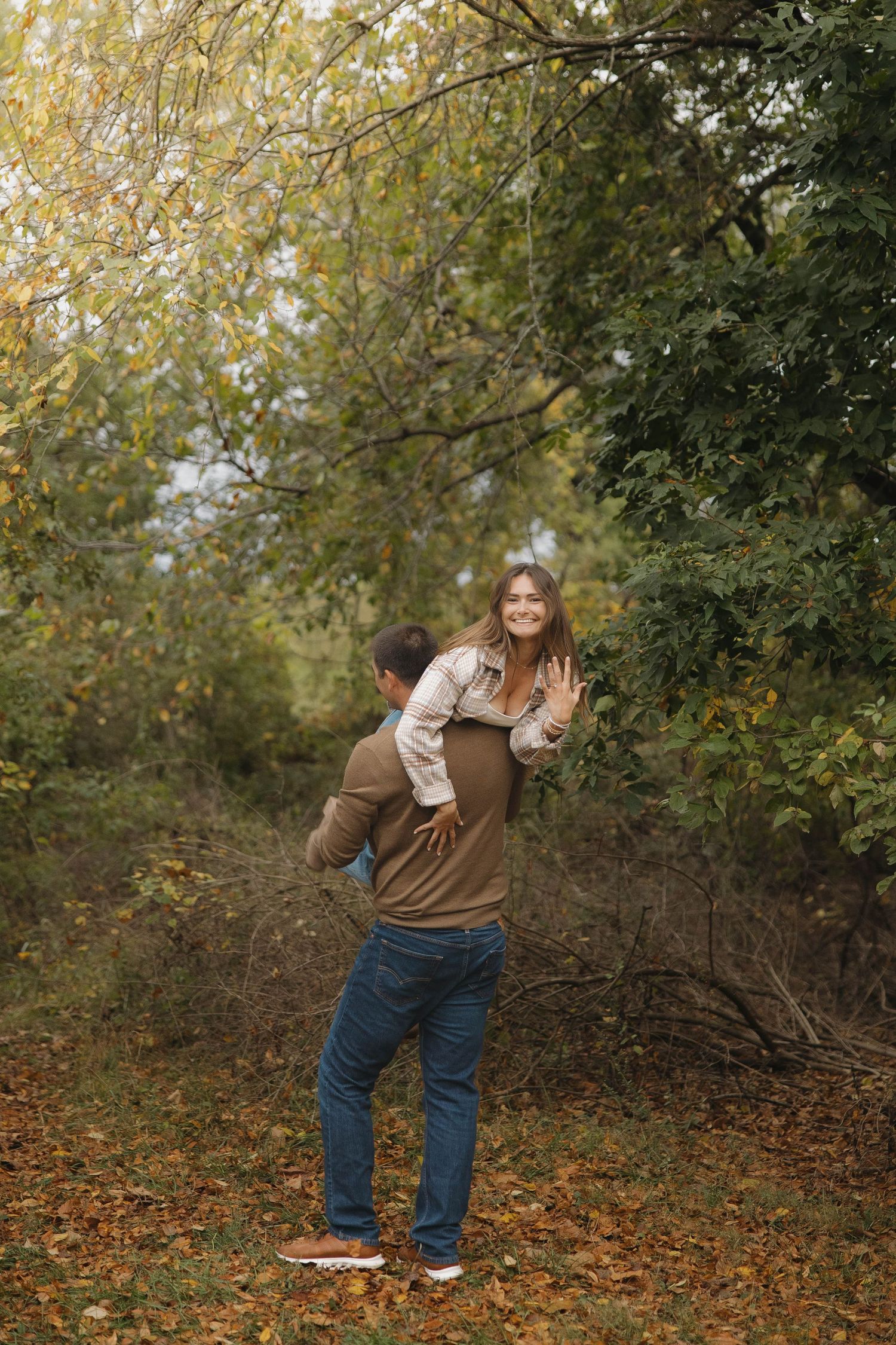 Person in brown sweater and jeans poses playfully among autumn trees with fallen leaves on the ground.