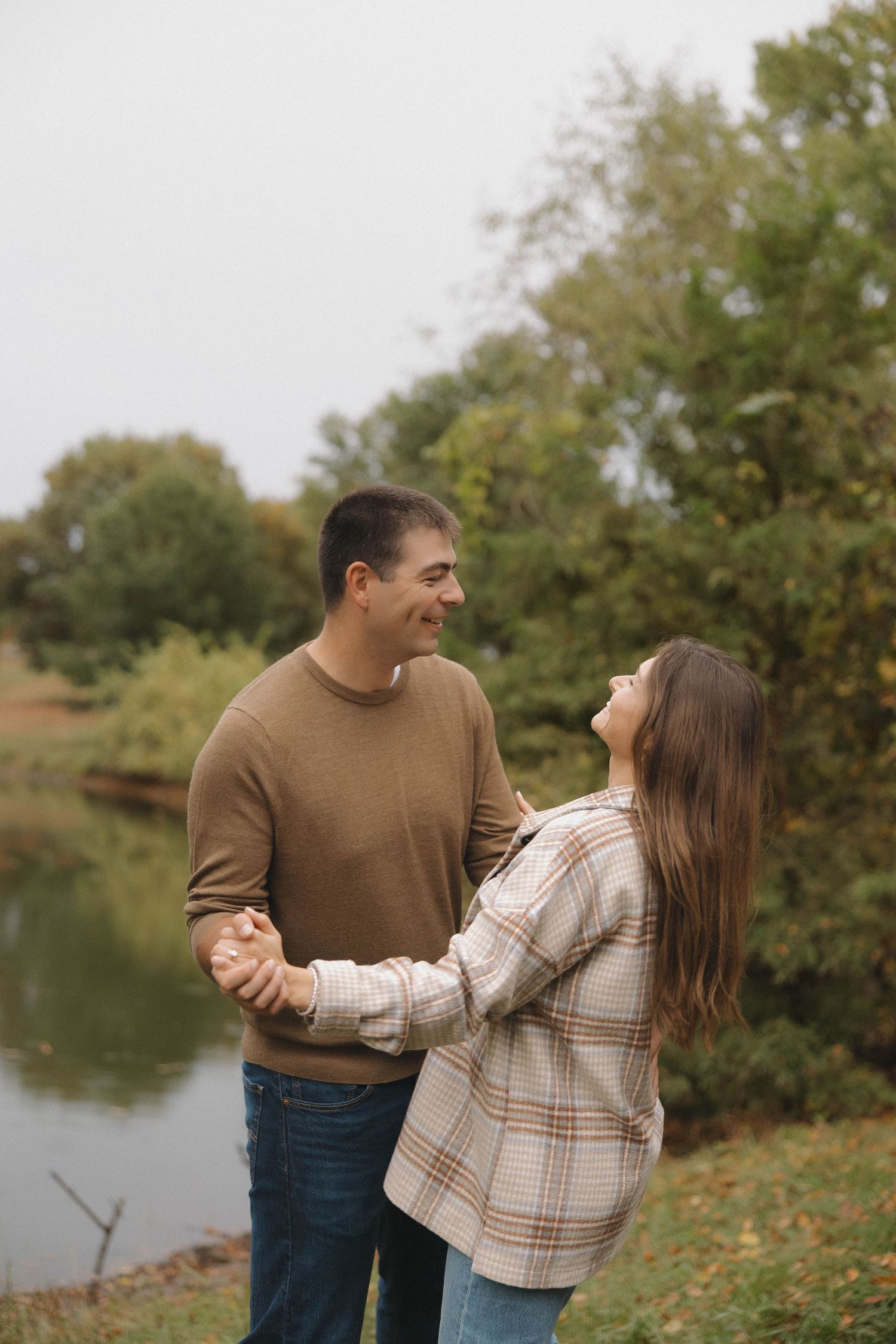 A couple shares a romantic moment while holding hands by a lake on an autumn day with trees in the background.