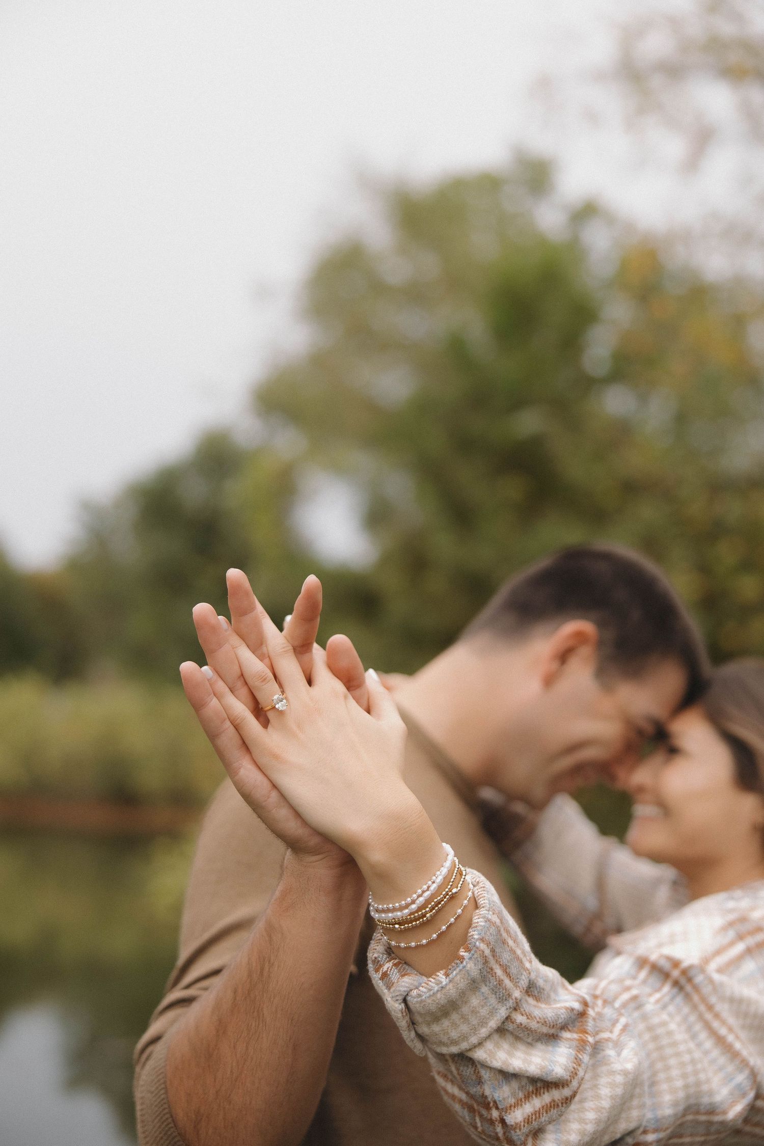 Two people hold hands while sitting outdoors near a lake with autumn trees in the background.