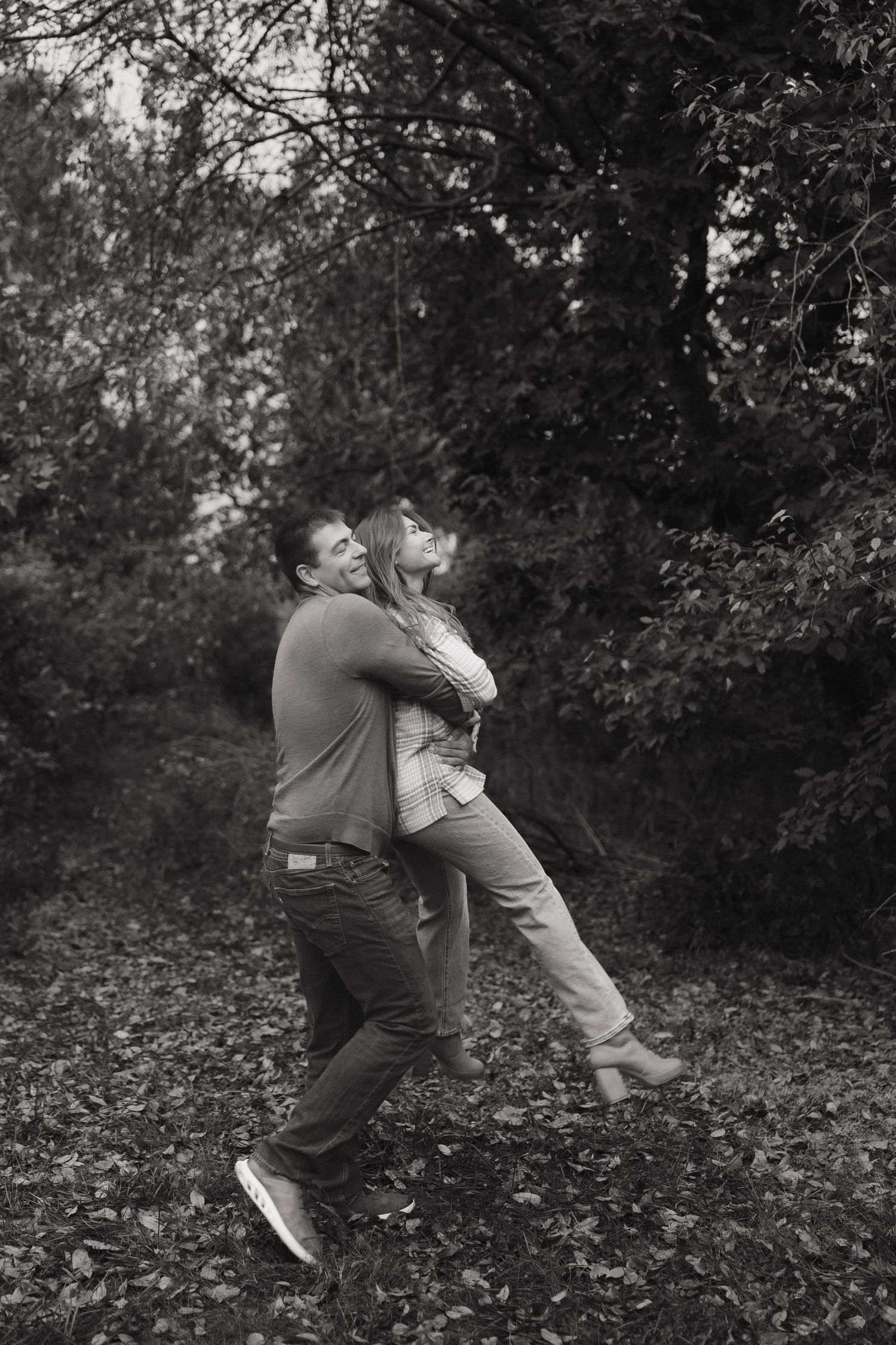 Black and white photo of a couple embracing in a leaf-covered forest setting.