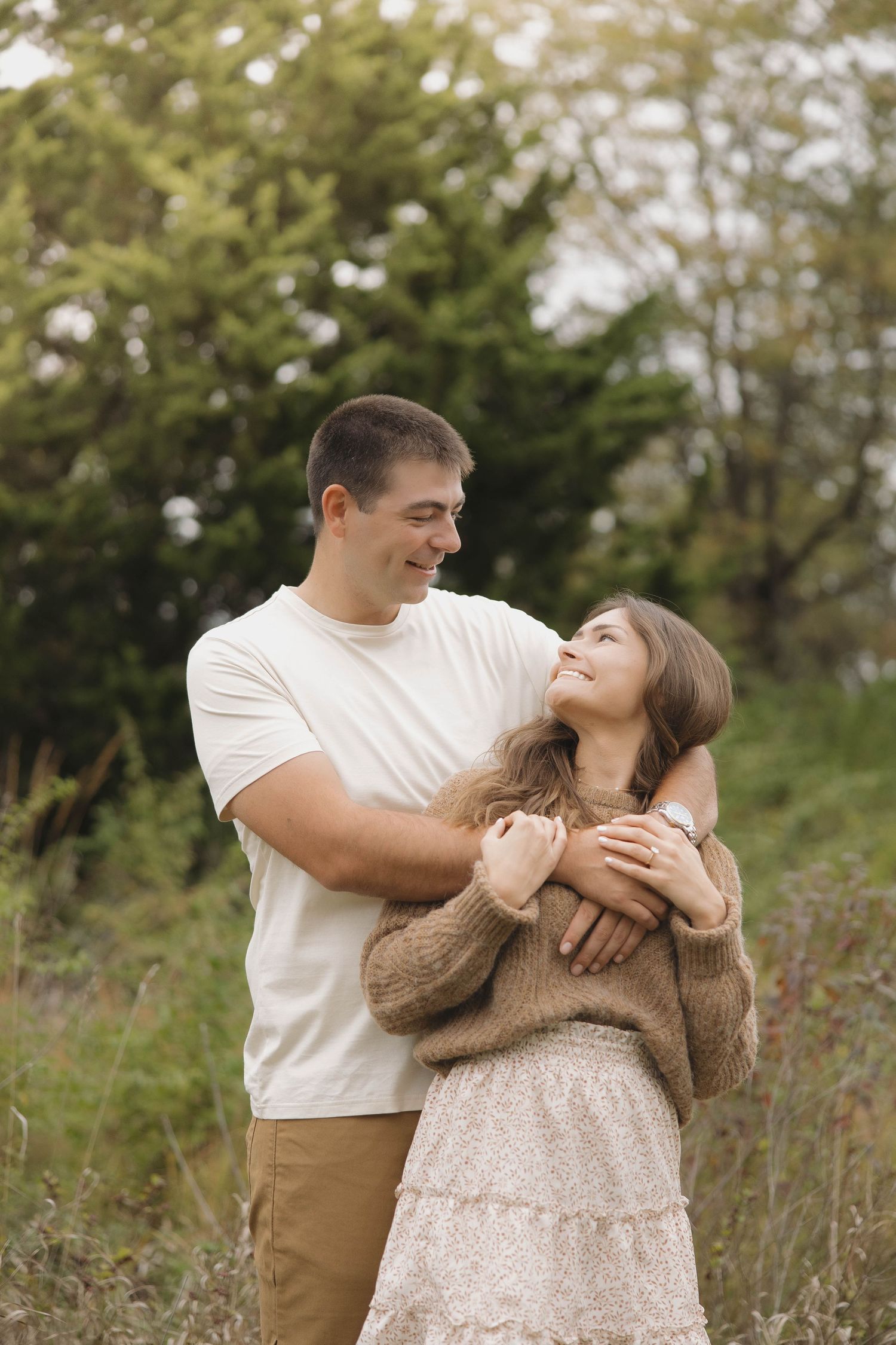 A romantic couple embraces in a field with autumn foliage and evergreen trees in the background.