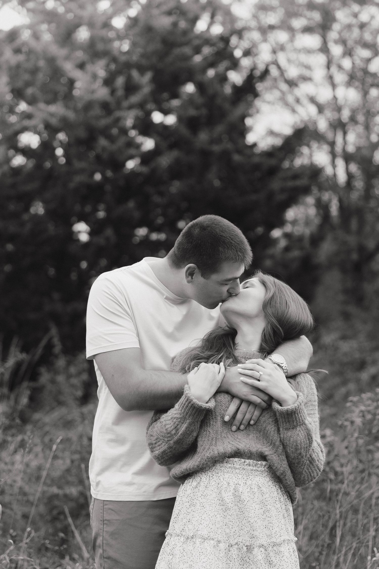 A romantic couple shares a tender kiss outdoors in a black and white photo with trees in the background.