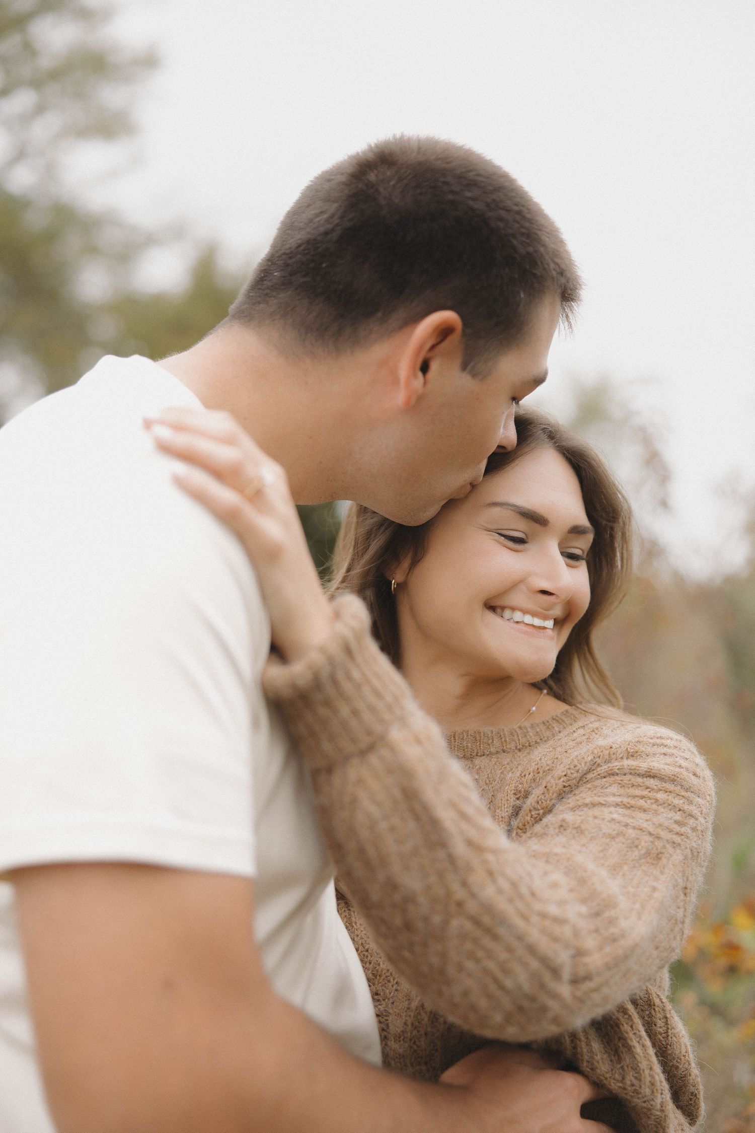 A tender romantic moment captured outdoors as a couple in casual attire embrace with genuine smiles.