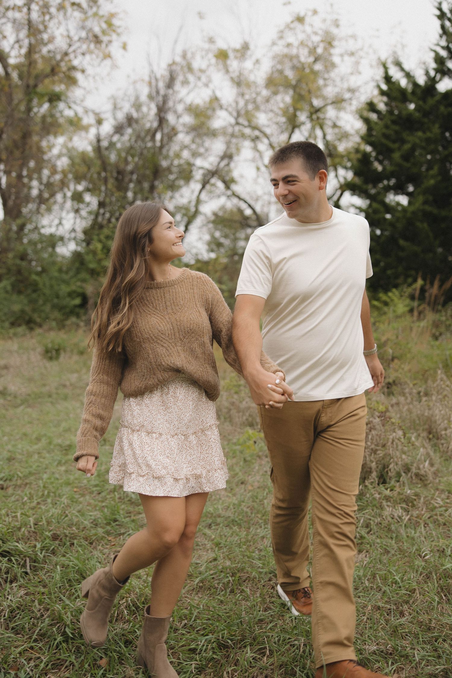 A couple walks hand in hand through a grassy field on an autumn day, wearing casual sweater and khaki attire.