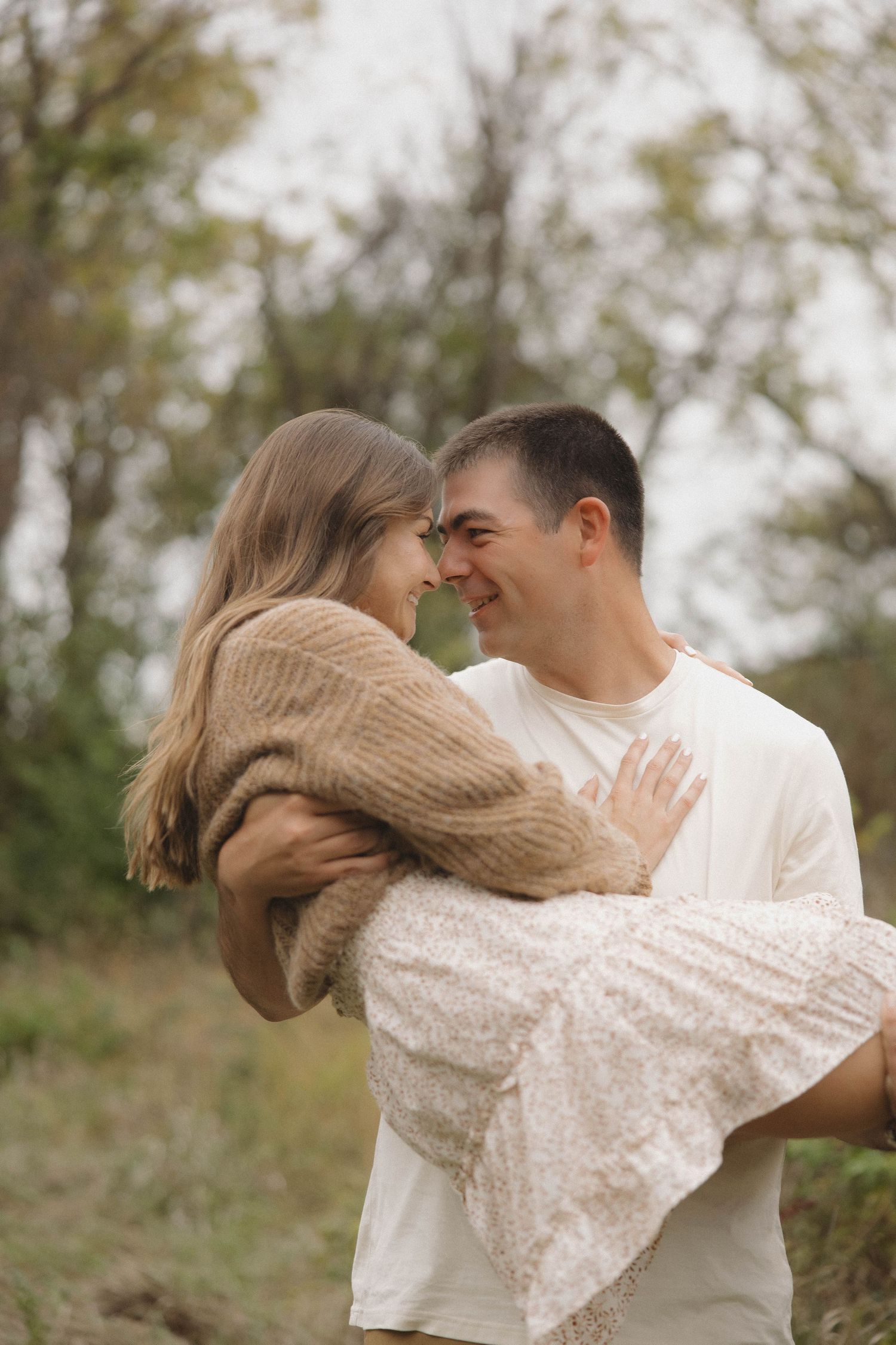 Joyful couple sharing an intimate moment outdoors, with her wearing a cozy brown sweater and white dress.