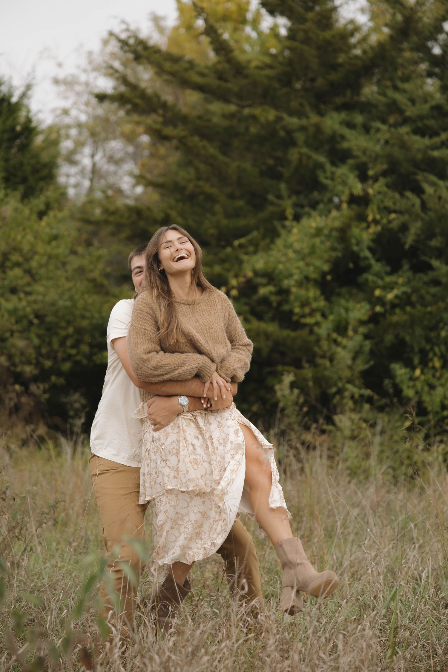 Couple sharing joyful moments together in a sunlit field with tall grass and evergreen trees in the background.