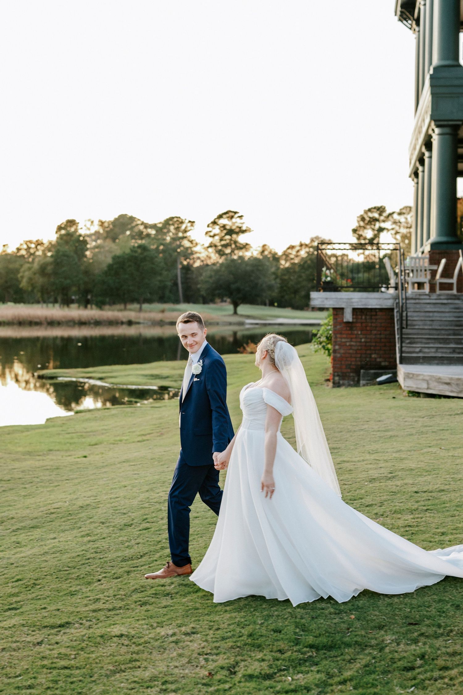 Newlyweds share a playful moment on a grassy lawn at sunset near a waterfront venue.