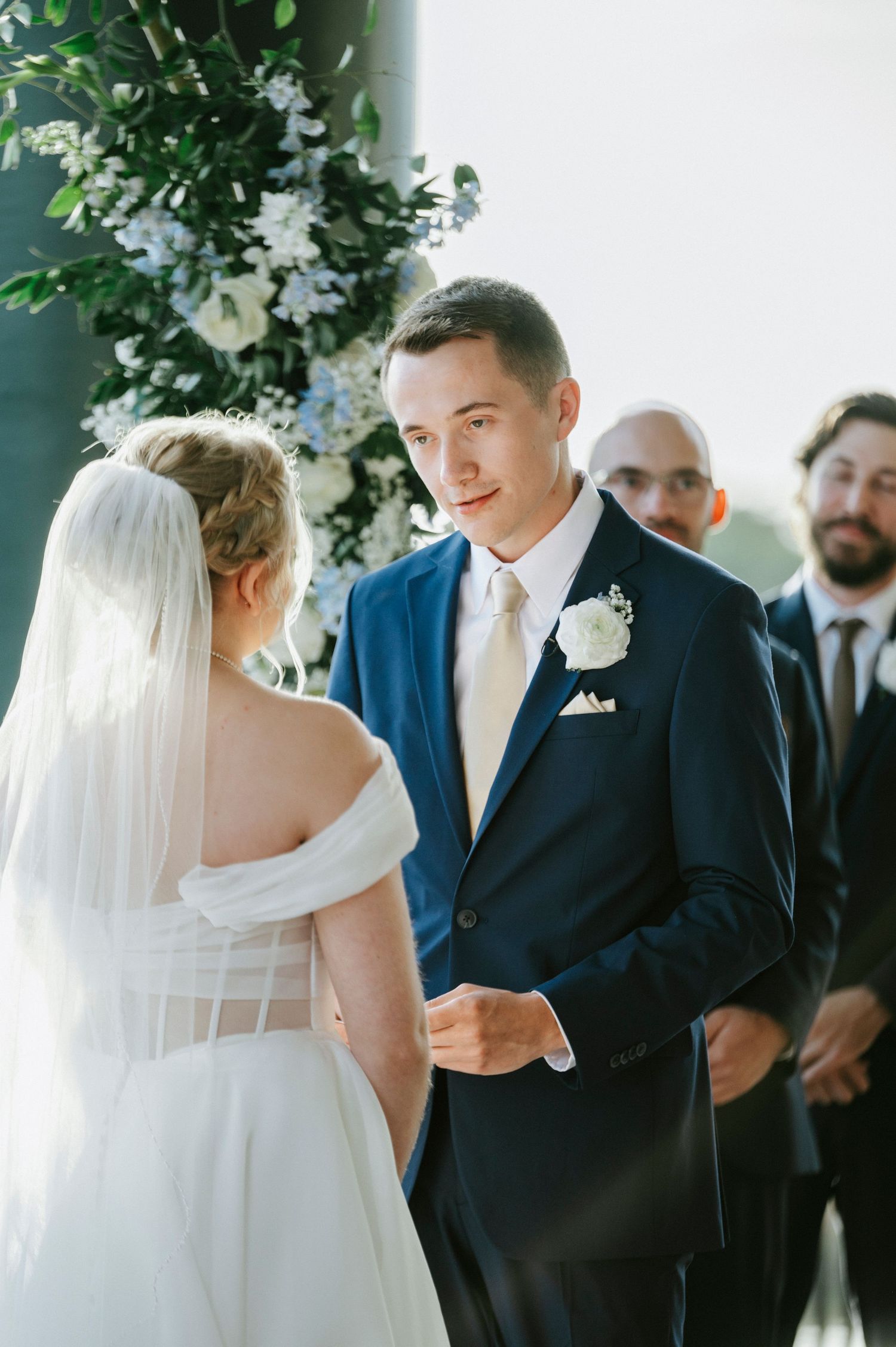 A wedding ceremony takes place under greenery with a couple exchanging vows while guests look on.