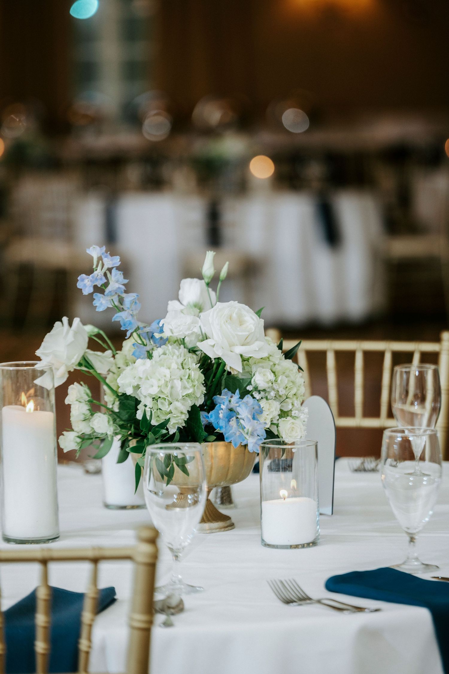 Elegant wedding reception table settings with white and blue floral centerpieces, candles, and gold chairs.