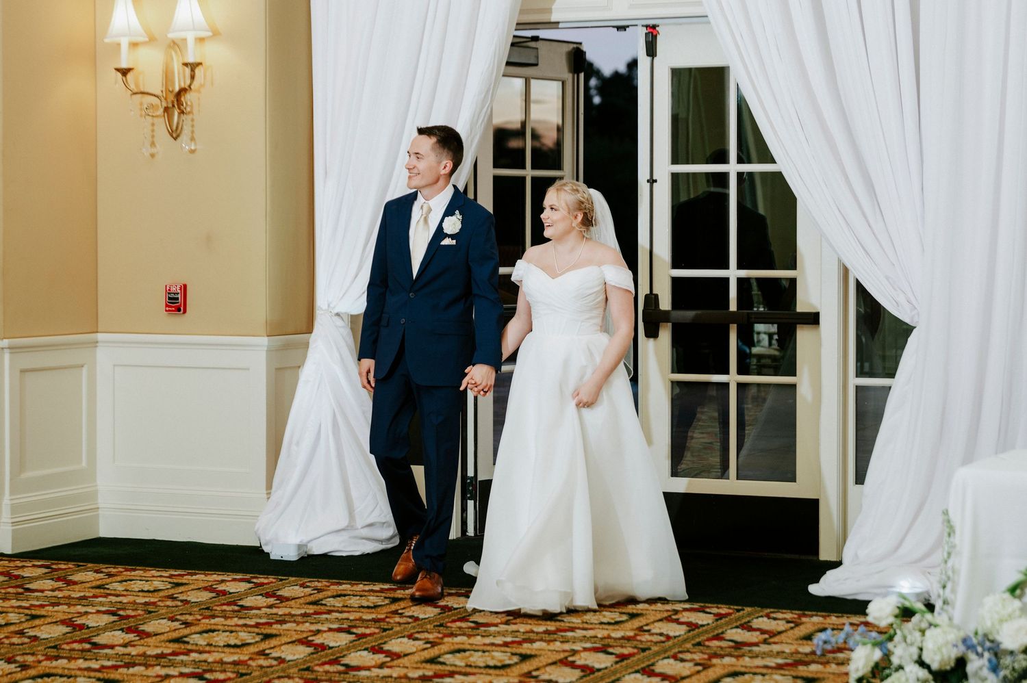 A romantic wedding scene with a couple in formal attire posing near white curtains in an elegant indoor venue.