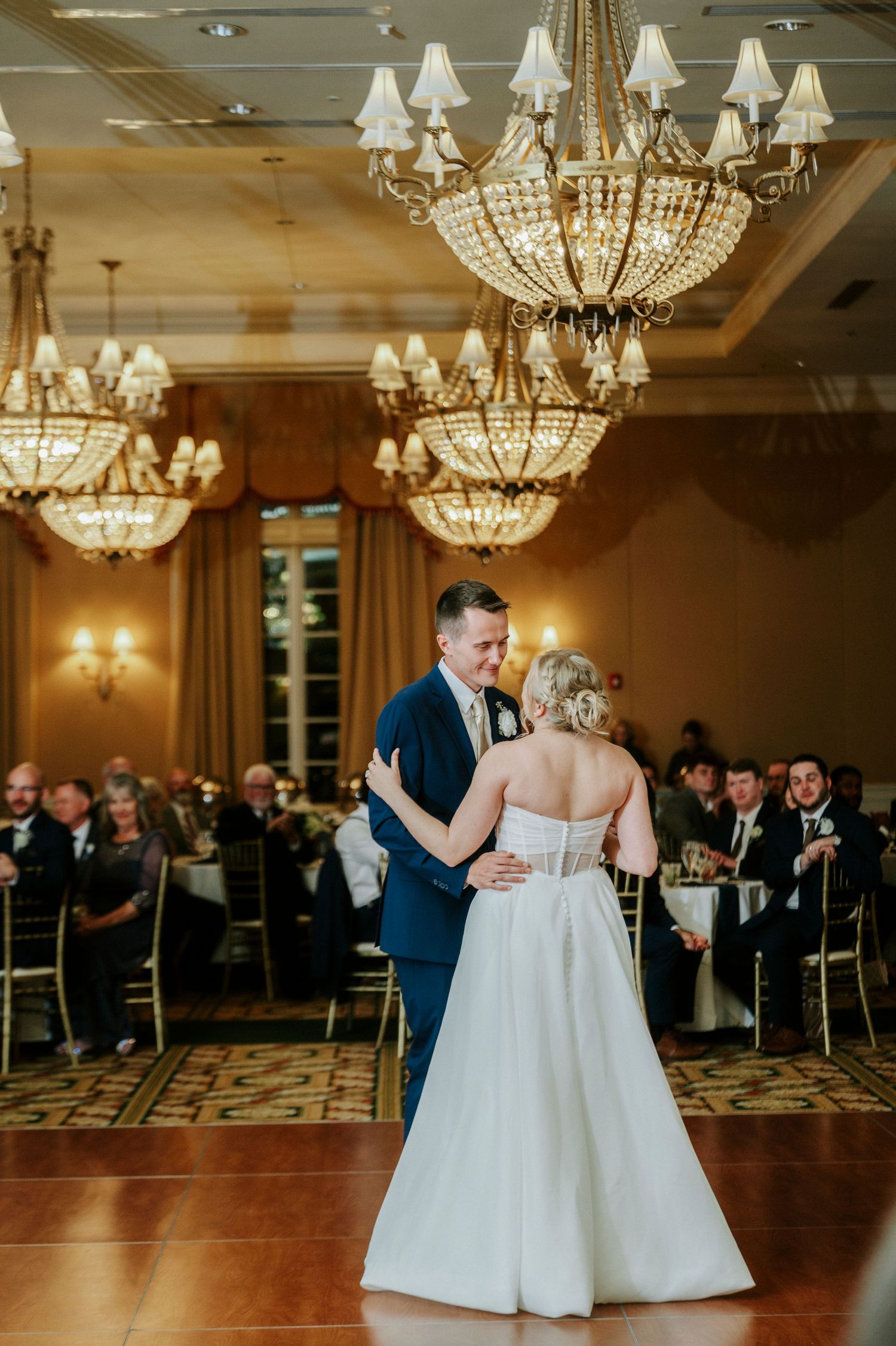 Wedding couple sharing their first dance on a wooden floor beneath ornate crystal chandeliers.