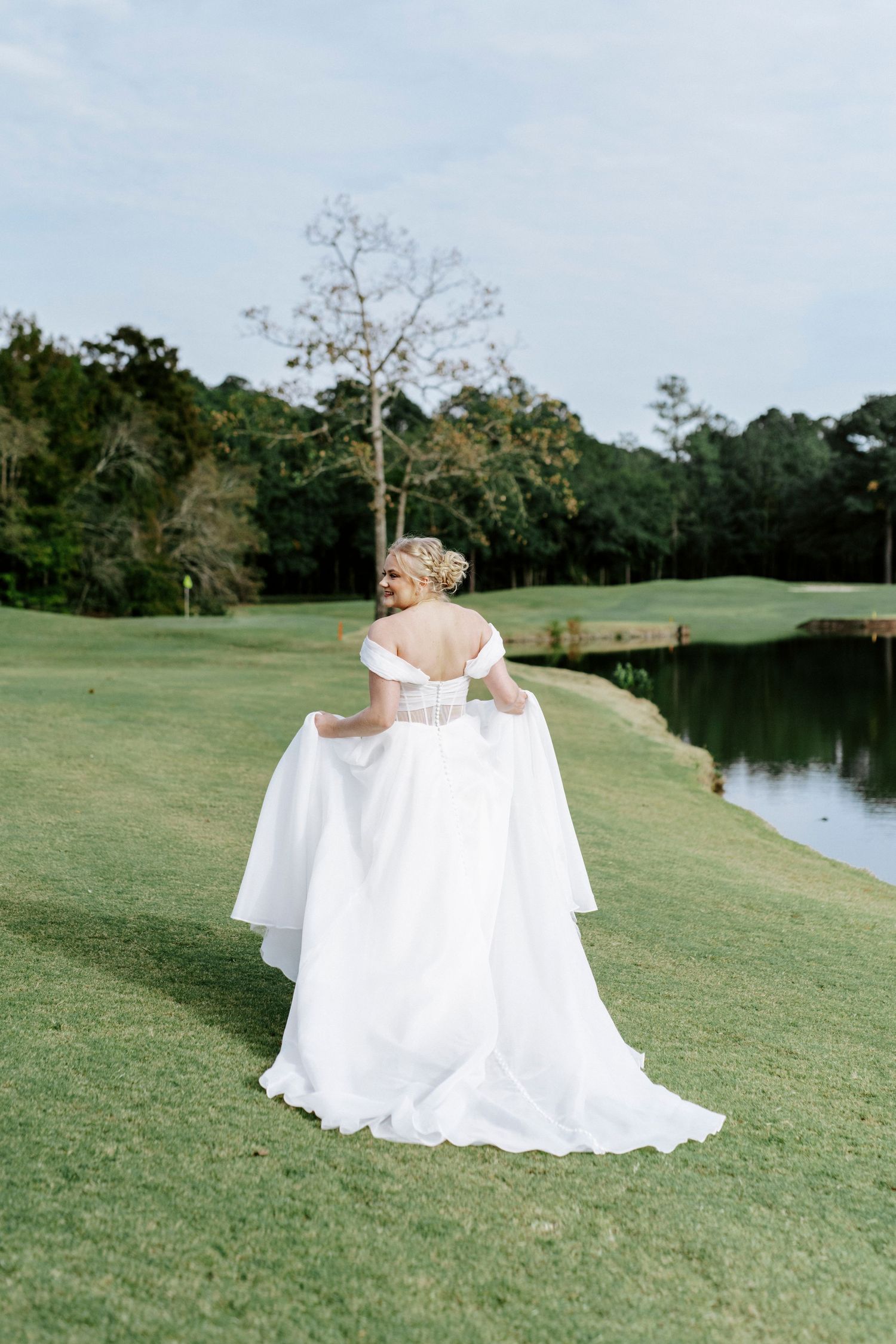 A bride in a flowing white gown twirls on a golf course beside a pond on her wedding day.