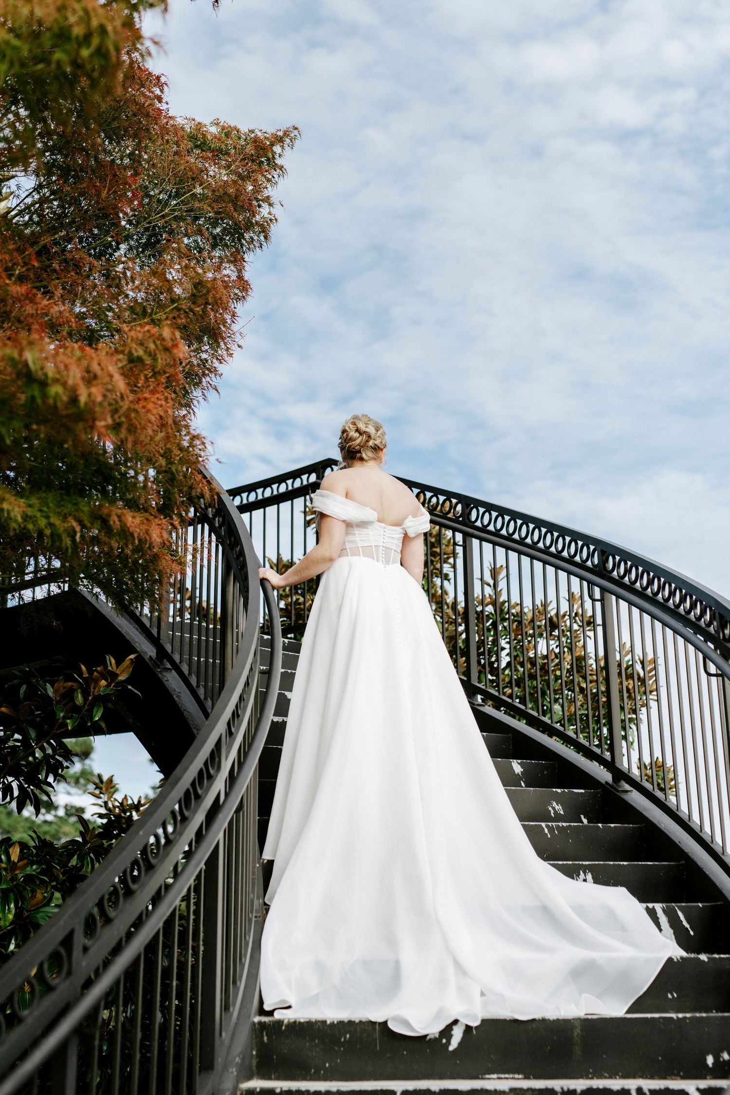 A bride in a flowing white gown stands atop an elegant curved black metal staircase with autumn foliage in the background.