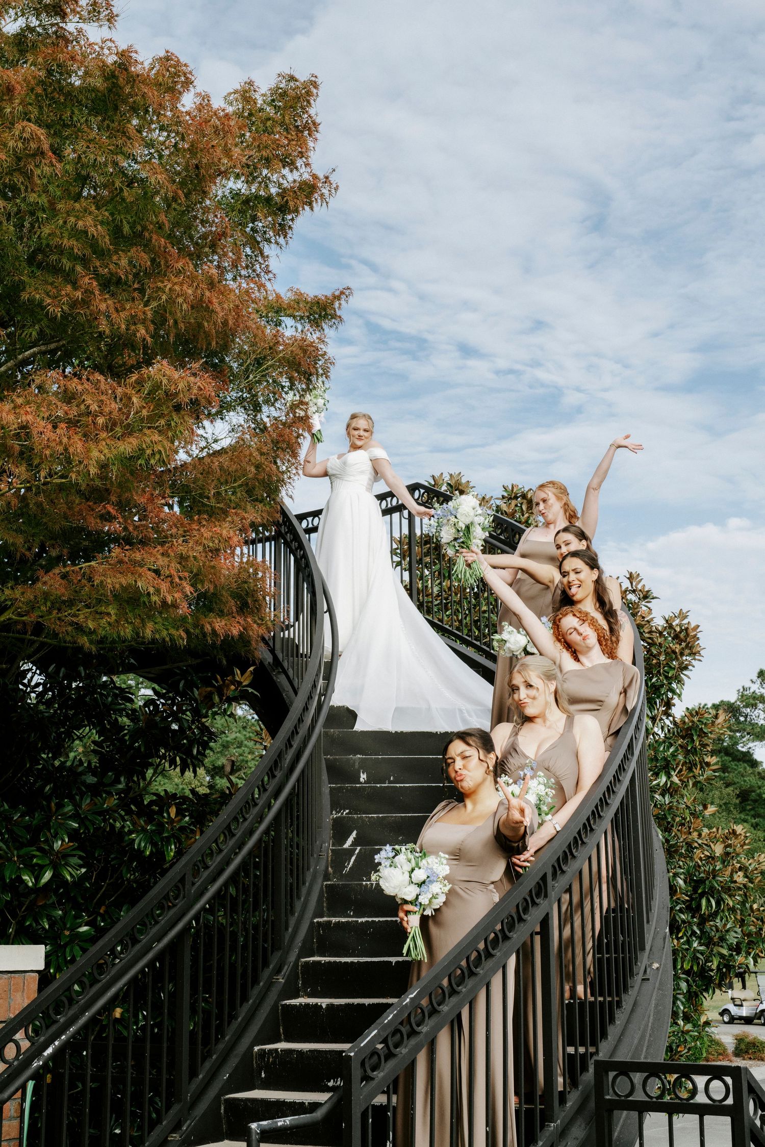 Wedding party poses playfully on outdoor stairs with autumn foliage in the background.