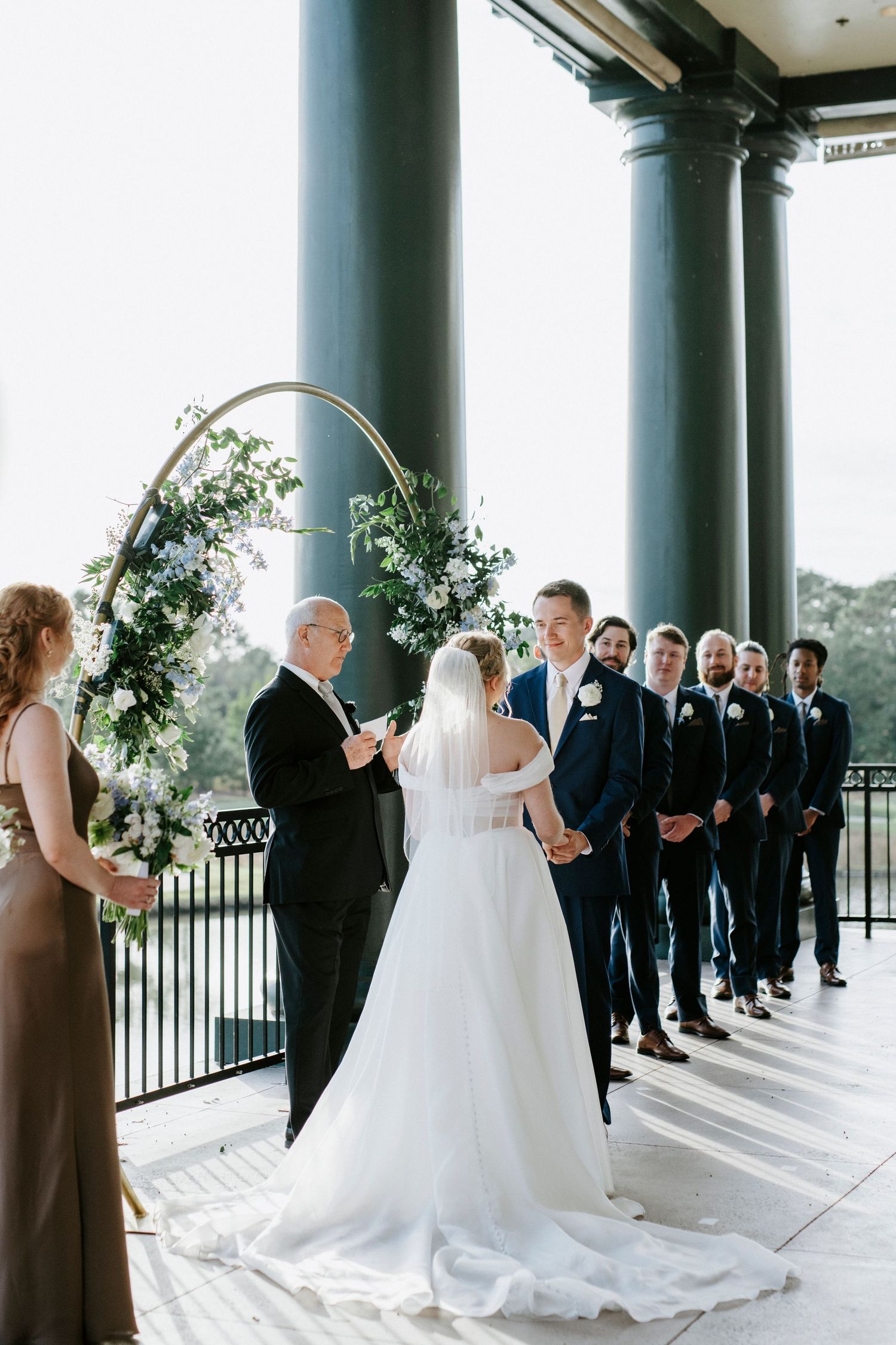 Wedding party lined up in formal attire under columned portico with flowing white gown.