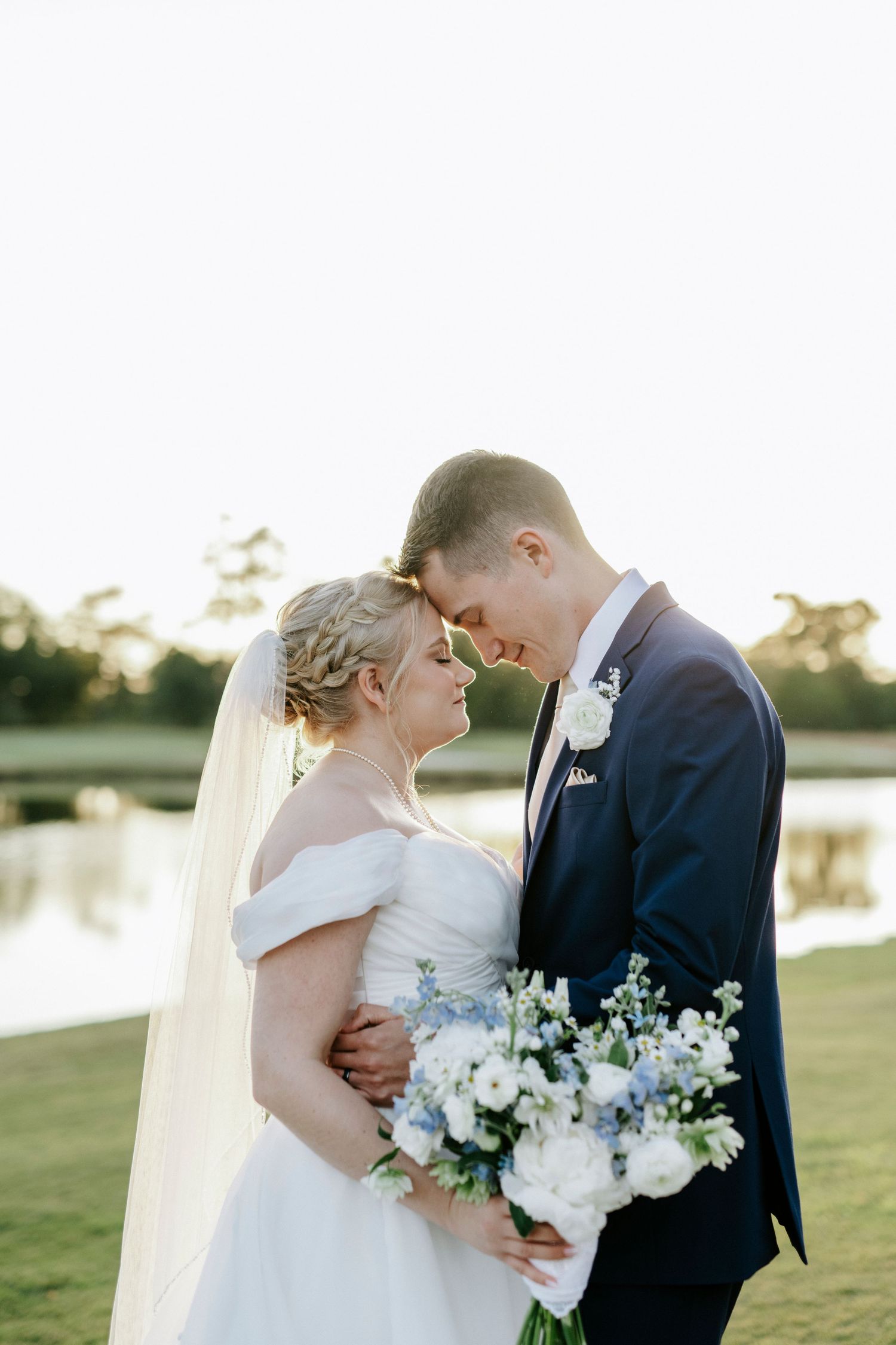 A bride and groom share romantic kisses at sunset by a lake while holding a white and blue floral bouquet.