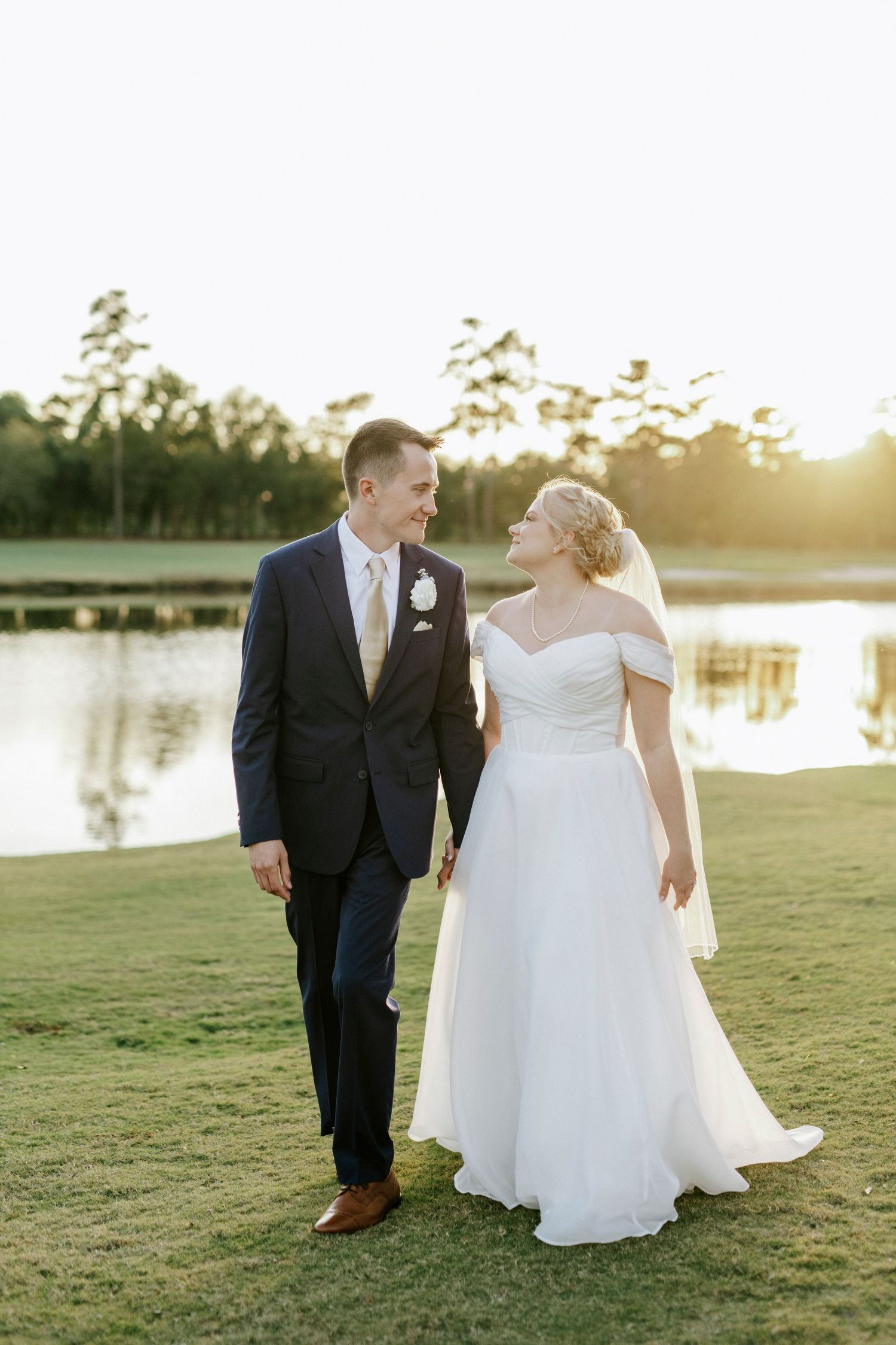 Newlyweds walking together on grassy lawn at sunset near a serene lake.