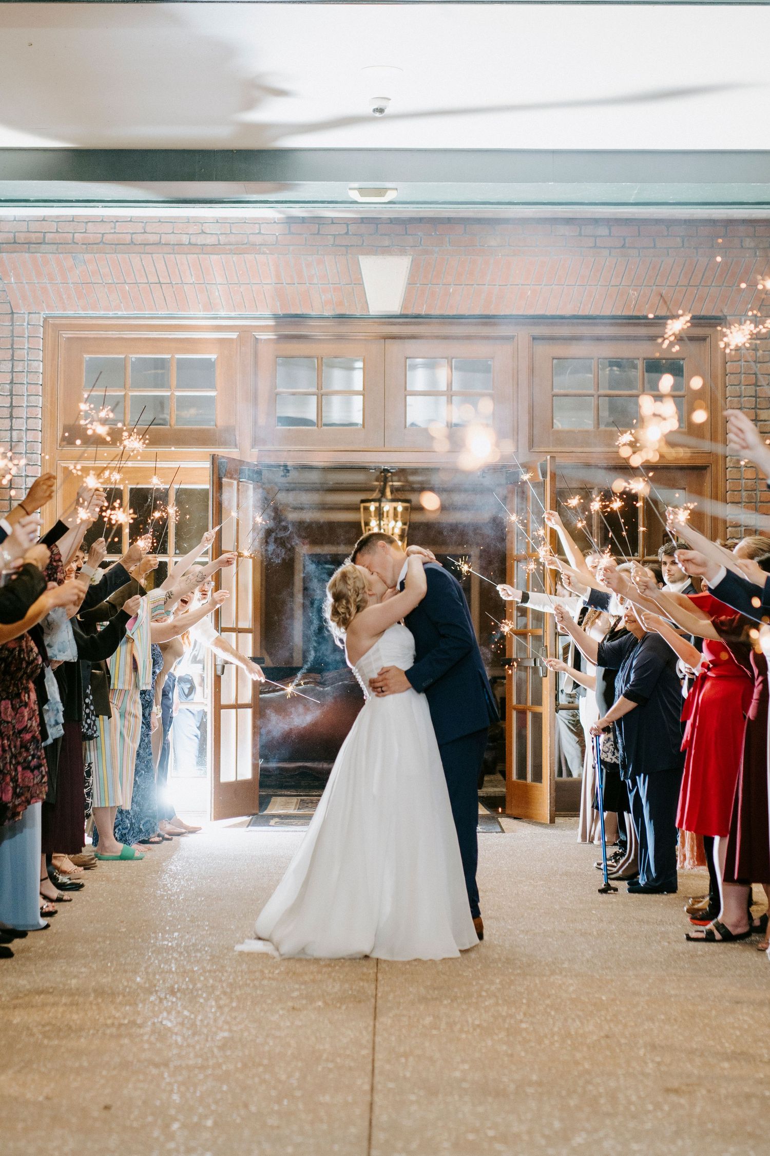 Wedding couple exits through a tunnel of guests waving sparklers in an indoor venue.