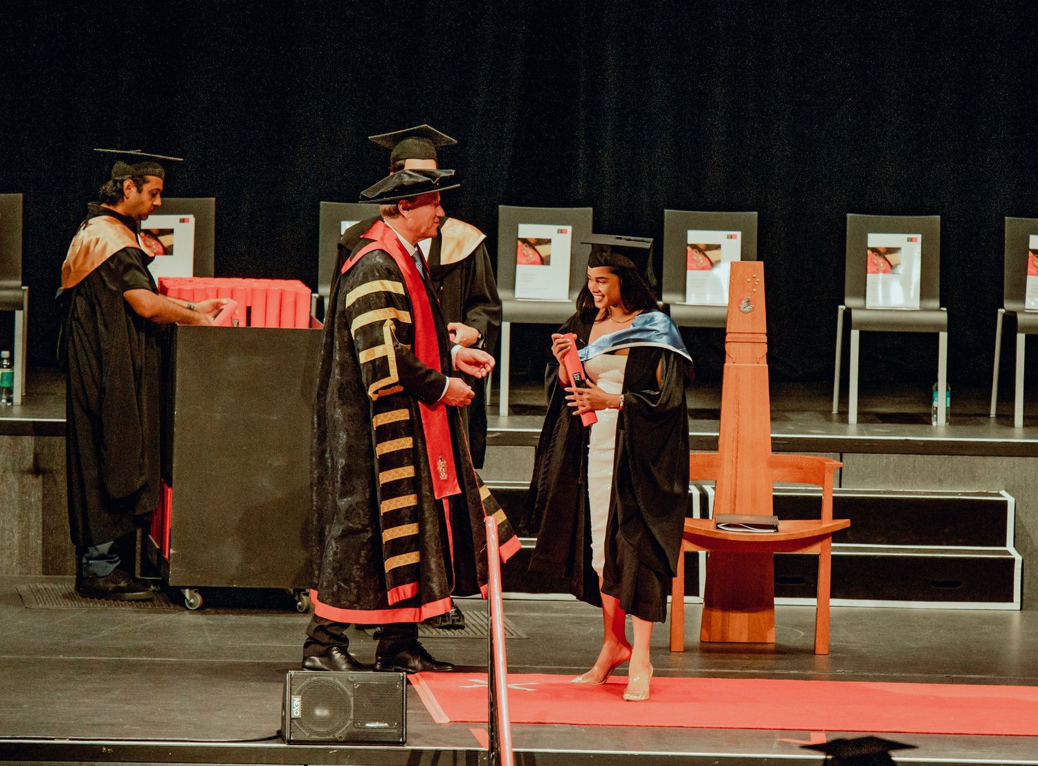 Graduates in red and black academic regalia walk across a stage during a university commencement ceremony.
