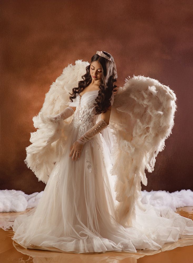 Ethereal maternity photoshoot featuring flowing white gown with dramatic angel wings against a dark brown backdrop.
