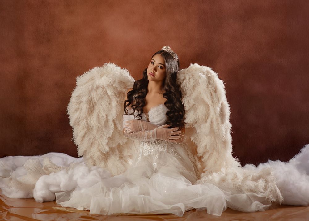 A figure in an ethereal white dress poses with large feathered angel wings against a dark brown backdrop.