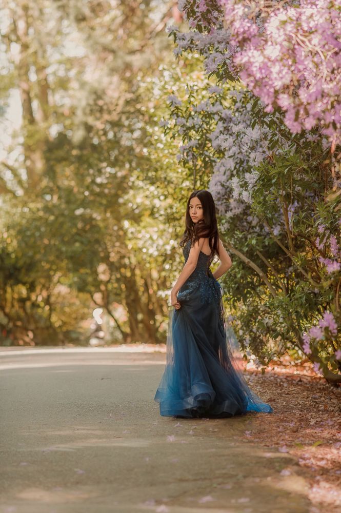 Woman in blue tulle gown poses gracefully among blooming purple flowers and sunlit trees in a dreamy outdoor setting.