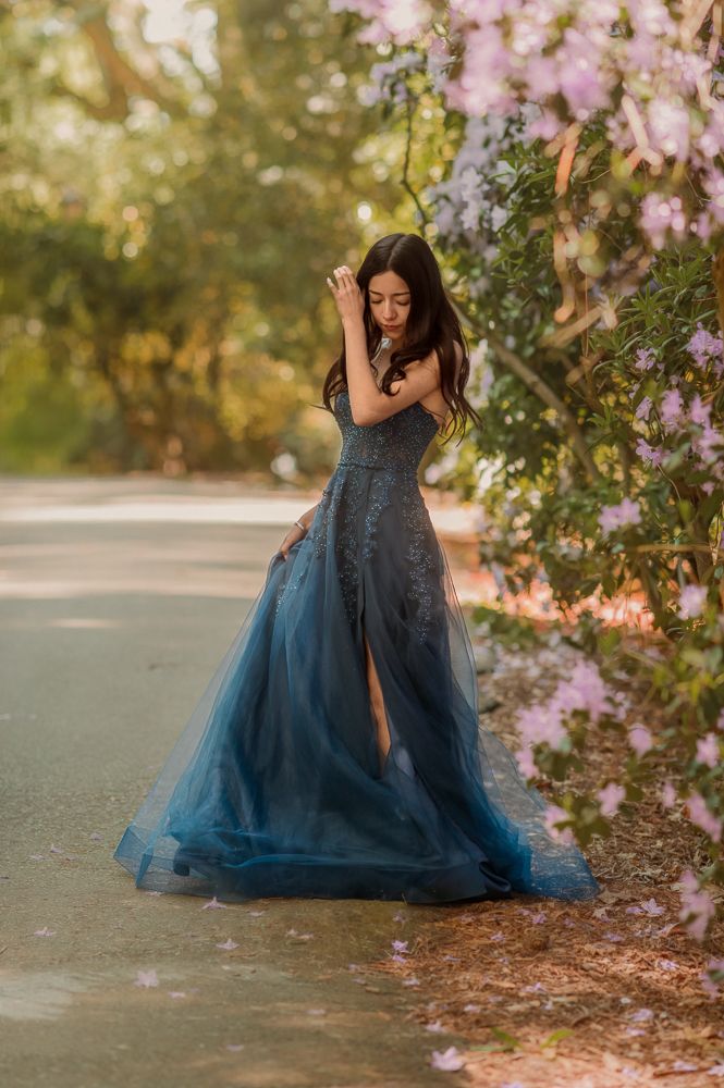 Woman in teal blue lace tulle gown poses near blooming pink flowers in a sunlit garden.