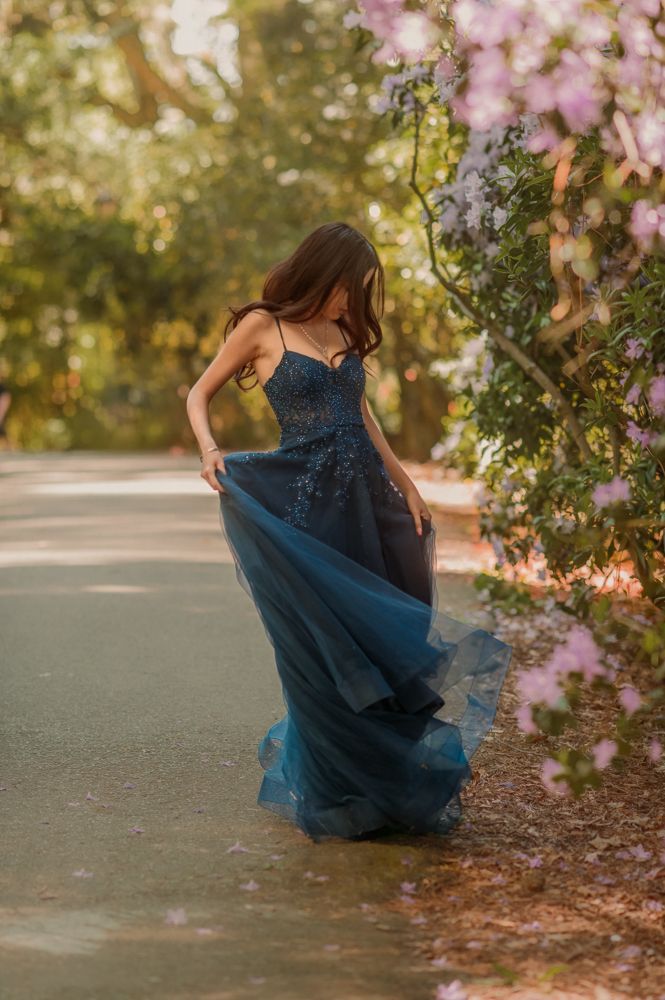 Woman in teal tulle gown poses among blooming pink flowers in golden hour light.
