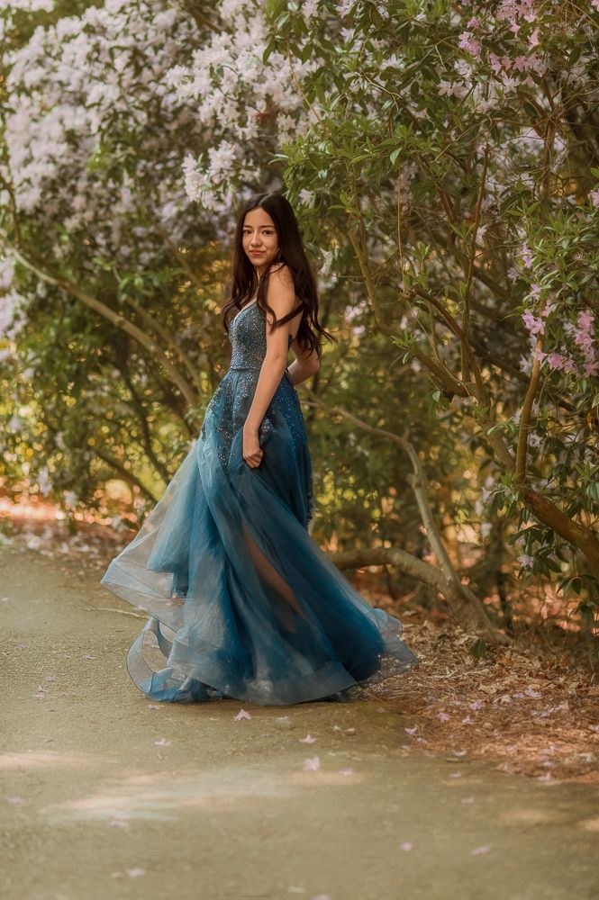Young woman in flowing teal tulle gown poses gracefully among blooming trees in a dreamy garden setting.