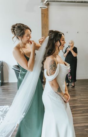 A bride in a white off-shoulder dress gets help with her veil from bridesmaid in sage green dress.