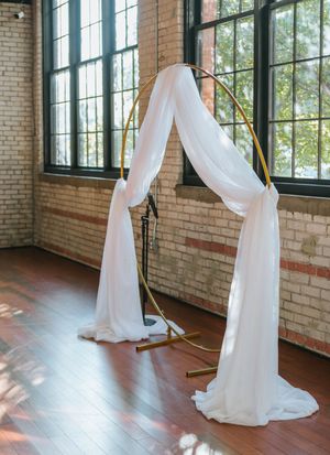 White fabric draped elegantly over a gold arch in an industrial brick-walled venue with large windows.